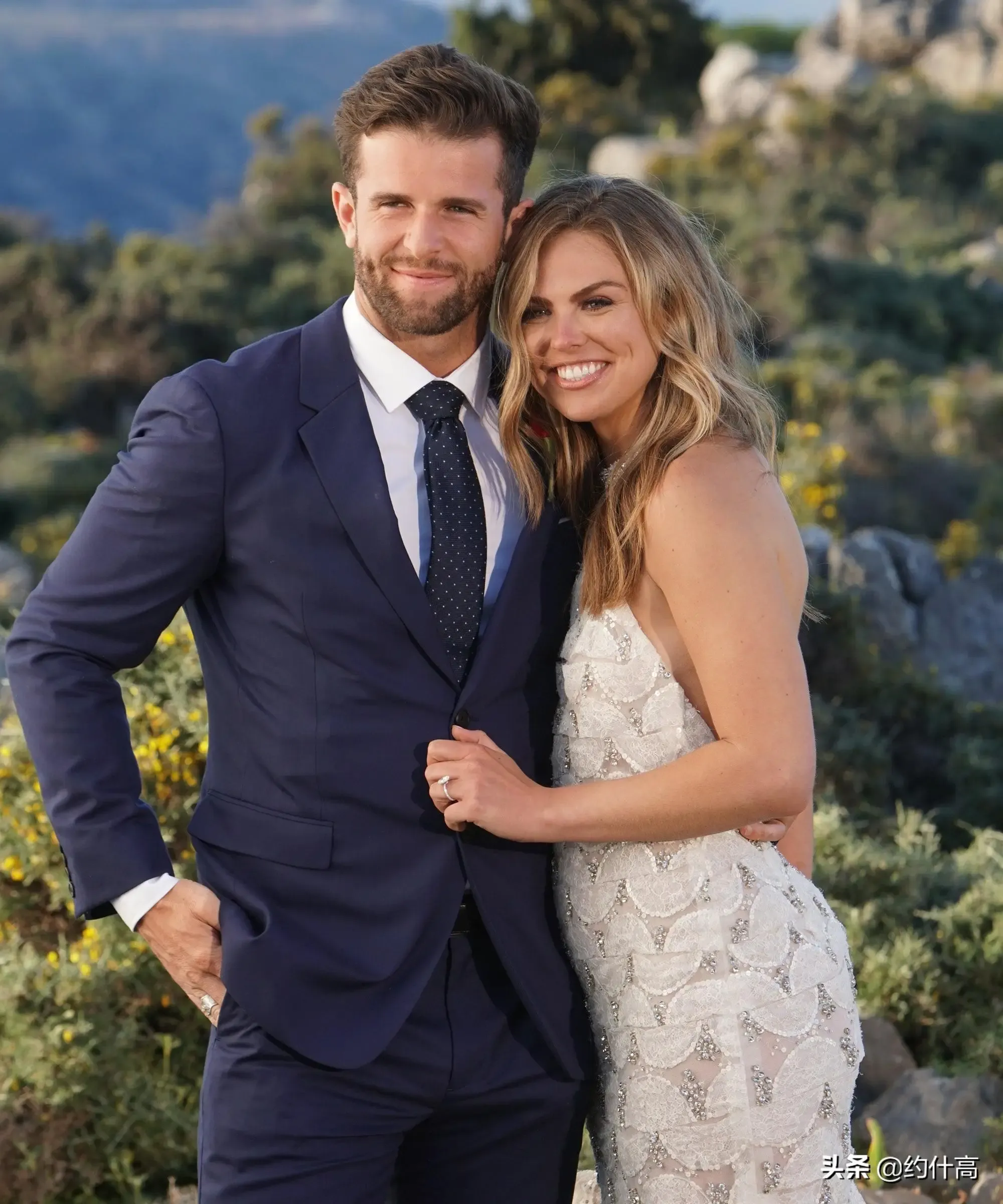 A wedding couple photo with the man in a suit and the woman in a white dress, standing in a lush, green outdoor setting.